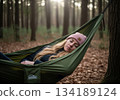 Woman relaxing and sleeping peacefully in a green hammock suspended between trees in a misty forest. 134189124