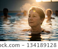 Serene senior woman enjoying a therapeutic hot spring bath with golden sunlight and steam 134189128