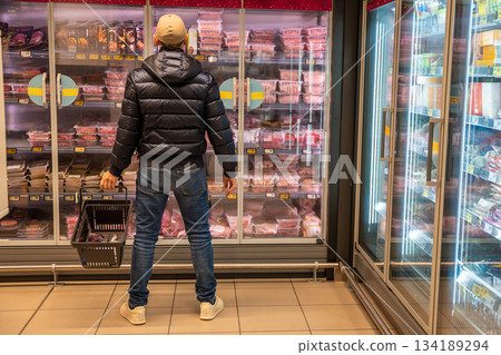 Man standing in front of a refrigerated meat section in a supermarket. Reflects everyday food choices and meat selection process during grocery shopping. 134189294
