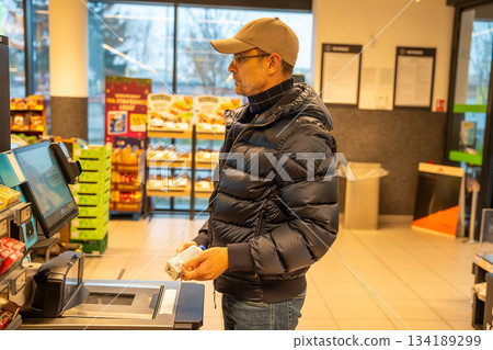 Man using a self-checkout machine at a grocery store, scanning items during a routine shopping trip. Concept of modern retail, automation, and independent shopping. 134189299