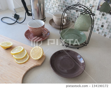 Ceramic cups and sliced lemon on wooden cutting board. Tea and cooking in kitchen. Food and drinks. Set of dishes on stand. 134190172