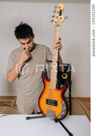 Vertical portrait of male musician, standing alongside bass guitar on stand, deeply concentrating and thinking about music composition, exploring inspiration, pursuing creative ideas in studio. 134191200