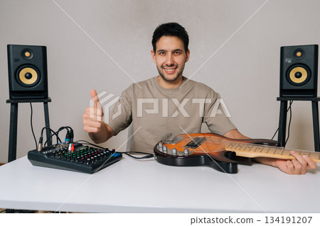 Happy excited man smiling and giving thumbs up, recording and producing music at home with professional equipment including audio mixer, bass guitar and studio monitors, copy space, isolated. Happy excited man smiling and giving thumbs up, recording and producing music at home with professional equipment including audio mixer, bass guitar and studio monitors, copy space, isolated. 134191207