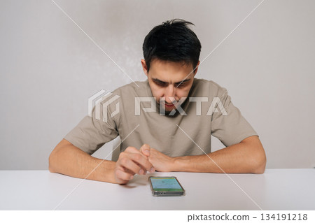 Portrait of young man sitting at white table, intently staring at smartphone with tense, focused expression while scrolling apps and searching for information indoors, white isolated background. Portrait of young man sitting at white table, intently staring at smartphone with tense, focused expression while scrolling apps and searching for information indoors, white isolated background. 134191218