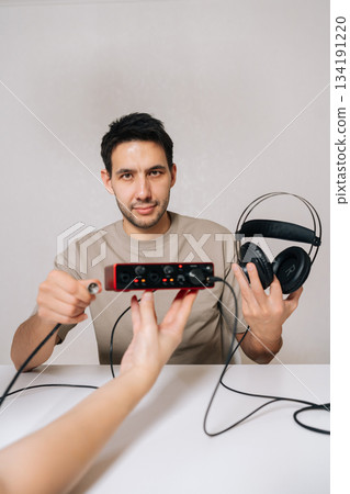Vertical portrait of man holding audio interface and headphones, demonstrating setup for sound recording or music production with hand offering cable connection sitting at white table looking camera. Vertical portrait of man holding audio interface and headphones, demonstrating setup for sound recording or music production with hand offering cable connection sitting at white table looking camera. 134191220