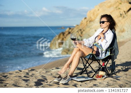 Naked businesswoman in shirt relaxing on sea beach with glass of red wine and picnic basket Naked businesswoman in shirt relaxing on sea beach with glass of red wine and picnic basket 134191727