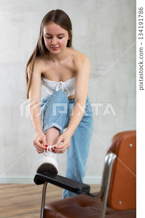 Girl with unfastened hair put foot on armrest of chair and tying shoelaces, lacing sneakers. 134191786