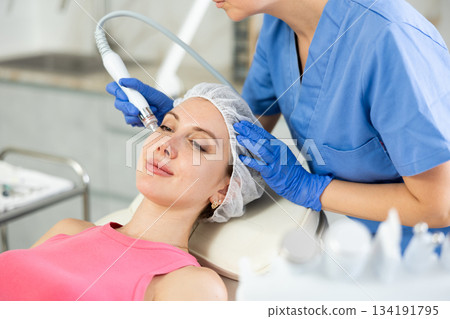 Young visitor receiving hardware electric massage and absorb blackheads and deeply clear skin by doctor in beauty clinic. Hardware cosmetology. Beautician hands in gloves makes face skin treatment for 134191795