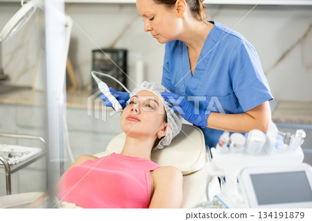 Experienced cosmetologist in rubber gloves does rf-lifting procedure on face for girl, smoothes wrinkles and tightens skin with use of electric hydrogen oxygen facial beauty machine in spa center Experienced cosmetologist in rubber gloves does rf-lifting procedure on face for girl, smoothes wrinkles and tightens skin with use of electric hydrogen oxygen facial beauty machine in spa center 134191879