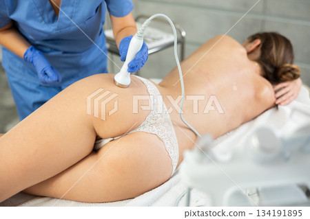 Adult woman cosmetologist gives ultrasonic massage of butt to young woman Adult woman cosmetologist gives ultrasonic massage of butt to young woman 134191895