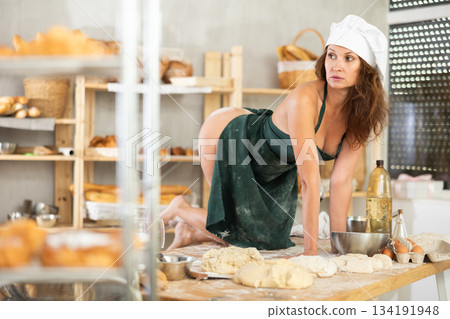 Sexy female baker in apron on naked body posing sexy on knees on kitchen table of bakery Sexy female baker in apron on naked body posing sexy on knees on kitchen table of bakery 134191948