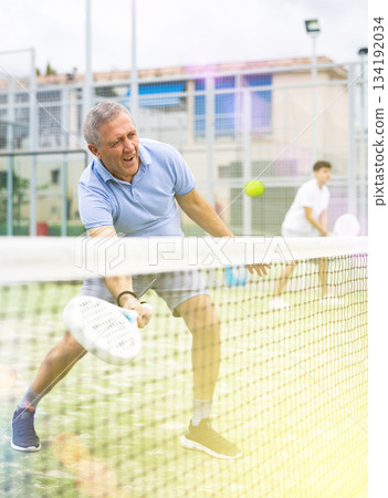 Positive elderly male player serving ball during training padel in court outdoors in spring 134192034