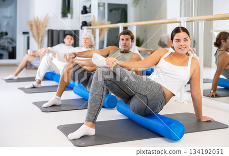 Young positive female performing set of exercises with pilates roller during group class in fitness studio. 134192051