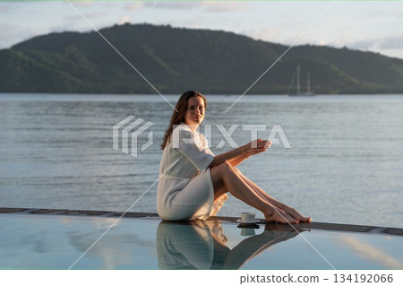 brunette white bathrobe sits at edge of pool overlooking Indian Ocean 134192066
