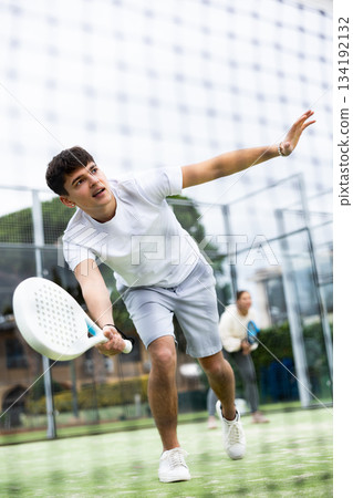Sportive young man playing padel with partner outdoors. View through tennis net 134192132