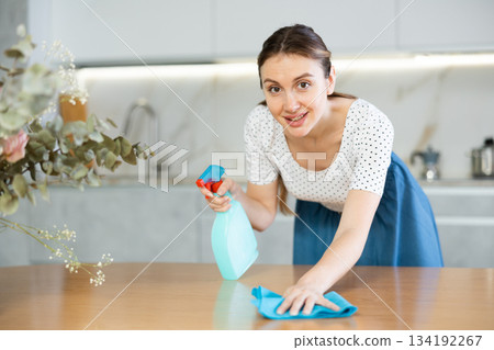 In kitchen, positive housewife in casual clothes holds rag in hands and cleans surface of table 134192267