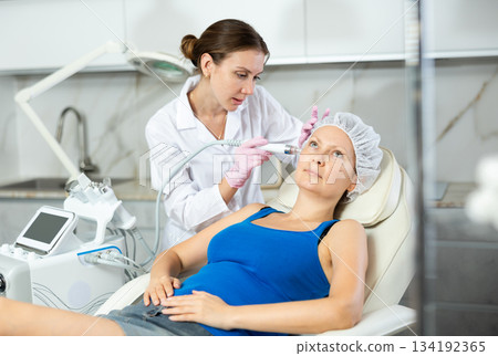 Professional female cosmetologist doing hydrafacial procedure in cosmetology clinic. Doctor use hydrogen and oxygen small bubbles for deeply clear skin. Rejuvenation and hydratation 134192365