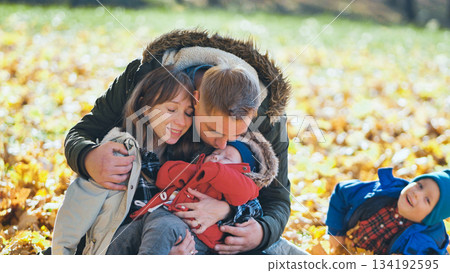 Parents cuddling their sleeping baby in a park full of fall foliage while their older son plays in the leaves 134192595