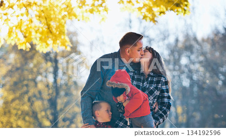 Parents kissing each other in a park during a beautiful autumn day, holding their two children, enjoying family life 134192596