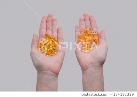 Hands holding two types of yellow softgel capsules containing D3 and Omega on a light  background. 134193389