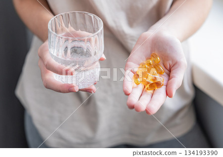 A close-up of a person's hands holding yellow softgels in one palm and a glass of water in the other. 134193390