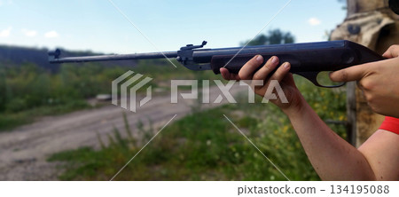 A person aims an air rifle outdoors, holding it steady with both hands. The background shows a dirt path and lush green field under a bright blue sky 134195088