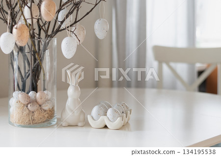 Decorative Easter tree with hanging speckled eggs and ceramic bunny on white table. Soft holiday decor in light pastel interior at kitchen. 134195538