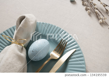 Elegant Easter table setting with blue speckled egg, gold cutlery and linen napkin on pastel plate and linen tablecloth. Soft spring decor and holiday atmosphere. Elegant Easter table setting with blue speckled egg, gold cutlery and linen napkin on pastel plate and linen tablecloth. Soft spring decor and holiday atmosphere. 134195539