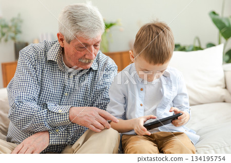 Happy family at home. Old senior man grandfather boy grandson enjoying time together watching video on phone. Good time at home. grandparent and child together. Older generation modern tech usage Happy family at home. Old senior man grandfather boy grandson enjoying time together watching video on phone. Good time at home. grandparent and child together. Older generation modern tech usage 134195754