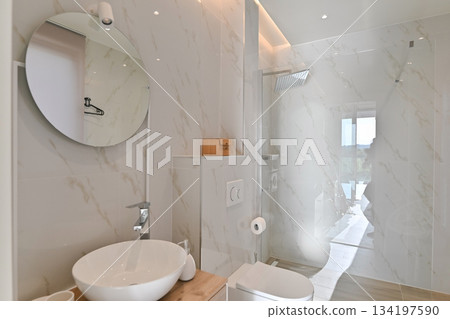 Modern, elegant bathroom featuring white marble-look tiles, a wooden vanity with a vessel sink, a wall-mounted toilet, and a glass shower, illuminated by sleek ambient lighting 134197590