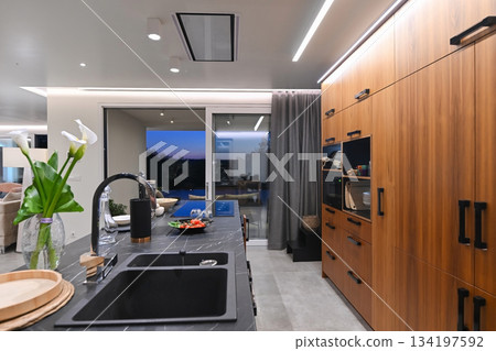 Modern luxury kitchen interior with wooden cabinetry, central island with bar stools, and ambient LED lighting. Stylish contemporary design with large windows and dark countertops at twilight 134197592