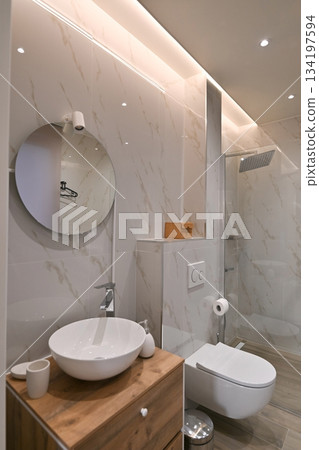 Modern, elegant bathroom featuring white marble-look tiles, a wooden vanity with a vessel sink, a wall-mounted toilet, and a glass shower, illuminated by sleek ambient lighting Modern, elegant bathroom featuring white marble-look tiles, a wooden vanity with a vessel sink, a wall-mounted toilet, and a glass shower, illuminated by sleek ambient lighting 134197594