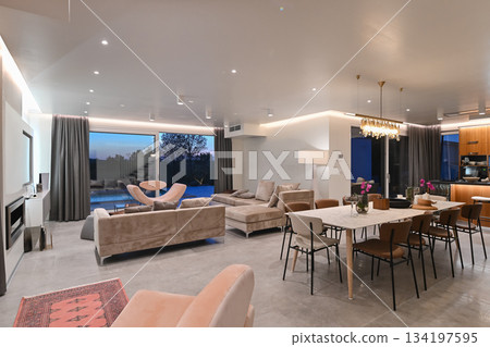 Modern luxury living room interior at sunset. Open-plan space featuring beige sofas, marble dining table, large glass windows with sun flare, and view of a patio. Stylish contemporary home design Modern luxury living room interior at sunset. Open-plan space featuring beige sofas, marble dining table, large glass windows with sun flare, and view of a patio. Stylish contemporary home design 134197595