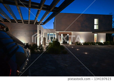 Exterior of a modern luxury house at night viewed from a carport. Illuminated white villa with paved driveway, wooden pergola, and car silhouette against a deep blue twilight sky 134197615