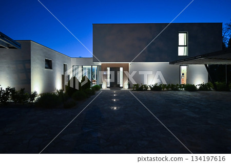 Exterior of a modern luxury house at night viewed from a carport. Illuminated white villa with paved driveway, wooden pergola, and car silhouette against a deep blue twilight sky 134197616