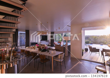 Modern luxury living room interior at sunset. Open-plan space featuring beige sofas, marble dining table, large glass windows with sun flare, and view of a patio. Stylish contemporary home design Modern luxury living room interior at sunset. Open-plan space featuring beige sofas, marble dining table, large glass windows with sun flare, and view of a patio. Stylish contemporary home design 134197619