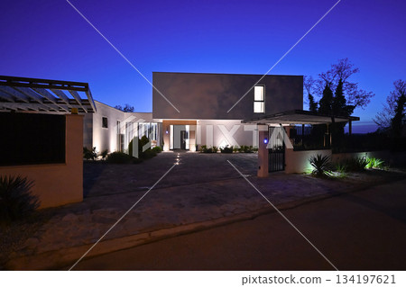 Exterior of a modern luxury house at night viewed from a carport. Illuminated white villa with paved driveway, wooden pergola, and car silhouette against a deep blue twilight sky 134197621