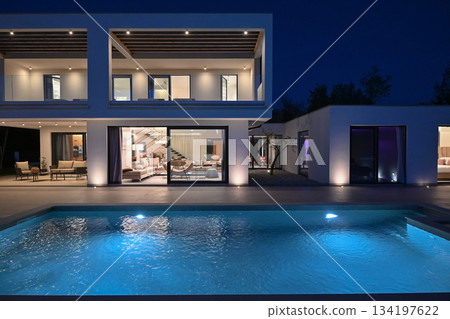 Exterior view of a modern luxury house with a swimming pool at twilight. Illuminated two-story white villa with large glass windows, patio, terrace, and glowing blue water 134197622