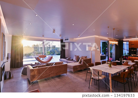 Modern luxury living room interior at sunset. Open-plan space featuring beige sofas, marble dining table, large glass windows with sun flare, and view of a patio. Stylish contemporary home design Modern luxury living room interior at sunset. Open-plan space featuring beige sofas, marble dining table, large glass windows with sun flare, and view of a patio. Stylish contemporary home design 134197633