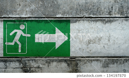 Green emergency exit sign with a running man and arrow, painted on a textured concrete wall. The sign indicates the direction of the nearest emergency exit route. Concept of Evacuation 134197870