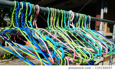 A Colorful Array of Plastic Clothes Hangers Hanging Outdoors on a Metal Rail in Sunlight 134198097