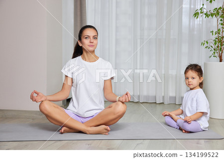 Mother and daughter sitting in lotus pose on yoga mats at home 134199522