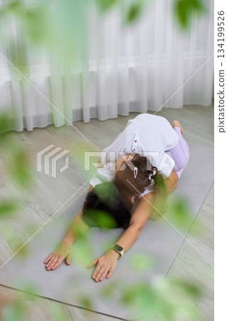Mother relaxing in child's pose while daughter claps hands, promoting family bonding and wellness Mother relaxing in child's pose while daughter claps hands, promoting family bonding and wellness 134199526