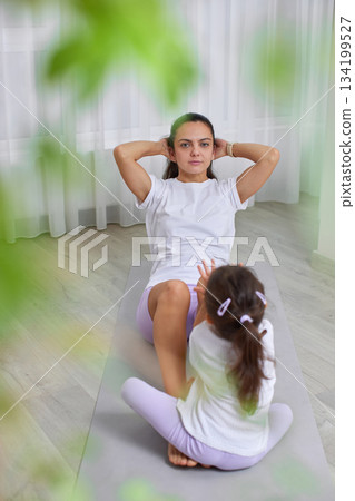 Mother doing crunches on a yoga mat with her daughter watching nearby Mother doing crunches on a yoga mat with her daughter watching nearby 134199527