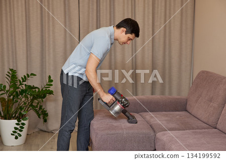 Man vacuuming sofa in living room, performing housework for maintaining home cleanliness 134199592