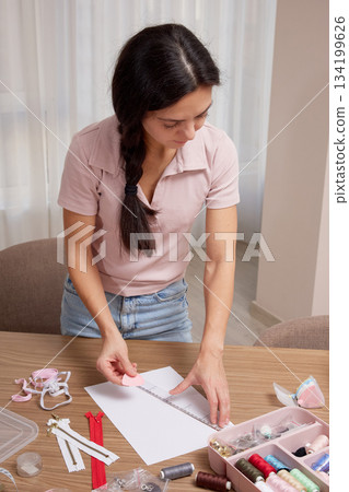 Woman focusing on drawing a fashion sketch at a table with sewing supplies Woman focusing on drawing a fashion sketch at a table with sewing supplies 134199626
