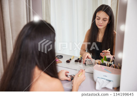 Woman holding cosmetic tube and brush, preparing makeup routine in front of mirror Woman holding cosmetic tube and brush, preparing makeup routine in front of mirror 134199645