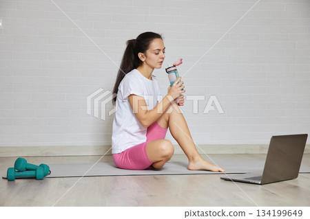 Young woman hydrating with a water bottle after exercising, using a laptop for online fitness Young woman hydrating with a water bottle after exercising, using a laptop for online fitness 134199649