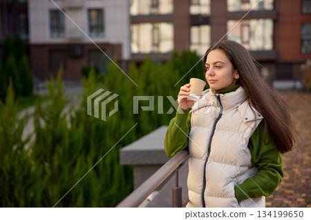 Woman standing on a modern park, drinking coffee and looking thoughtful 134199650