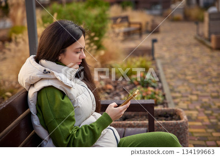 Woman sitting on a park bench, texting on her mobile phone 134199663